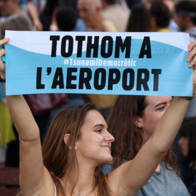 Pla d'una noia que mostra un cartell de Tsunami Democràtic cridant a la gent a mobilitzar-se a l'aeroport