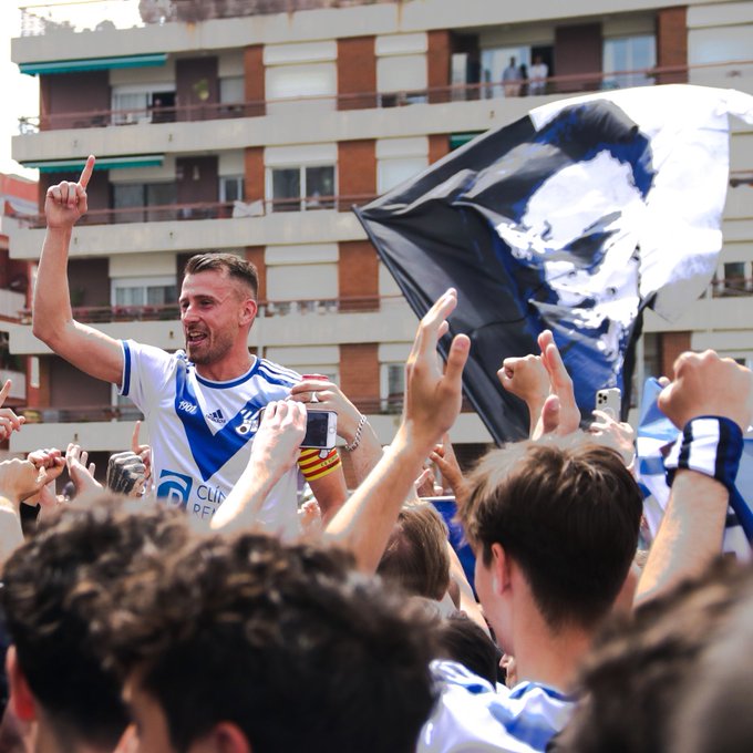 "El jugador i capità del CE Europa és aixecat pels seus companys, al seu darrera té una bandera amb la cara de Manuel Cros. Foto: Àngel Garreta". 